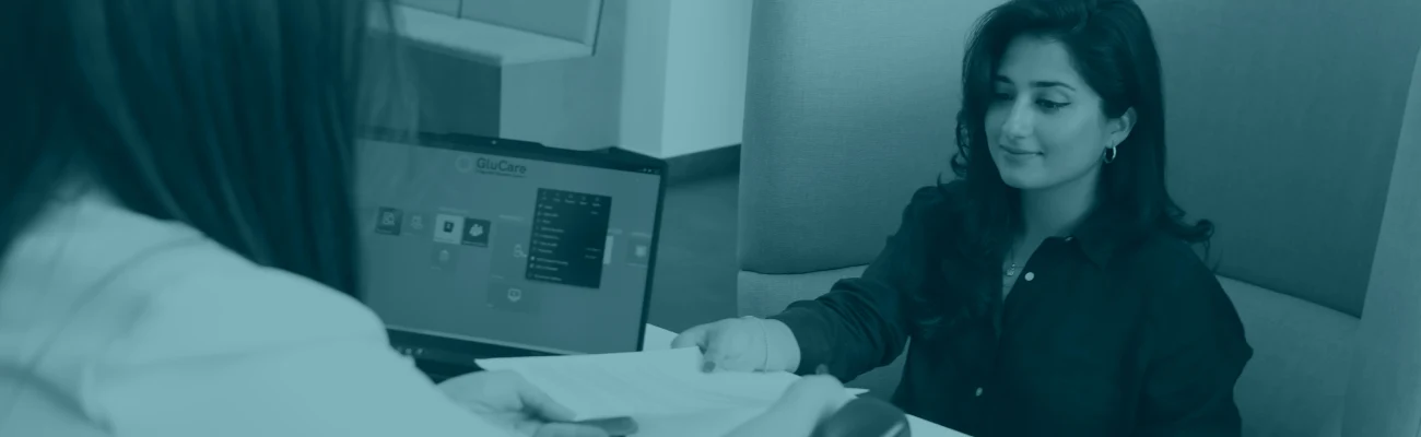 Teal-tinted banner image of a patient meeting with a healthcare professional discussing insurance and subscriptions, with medical device on desk